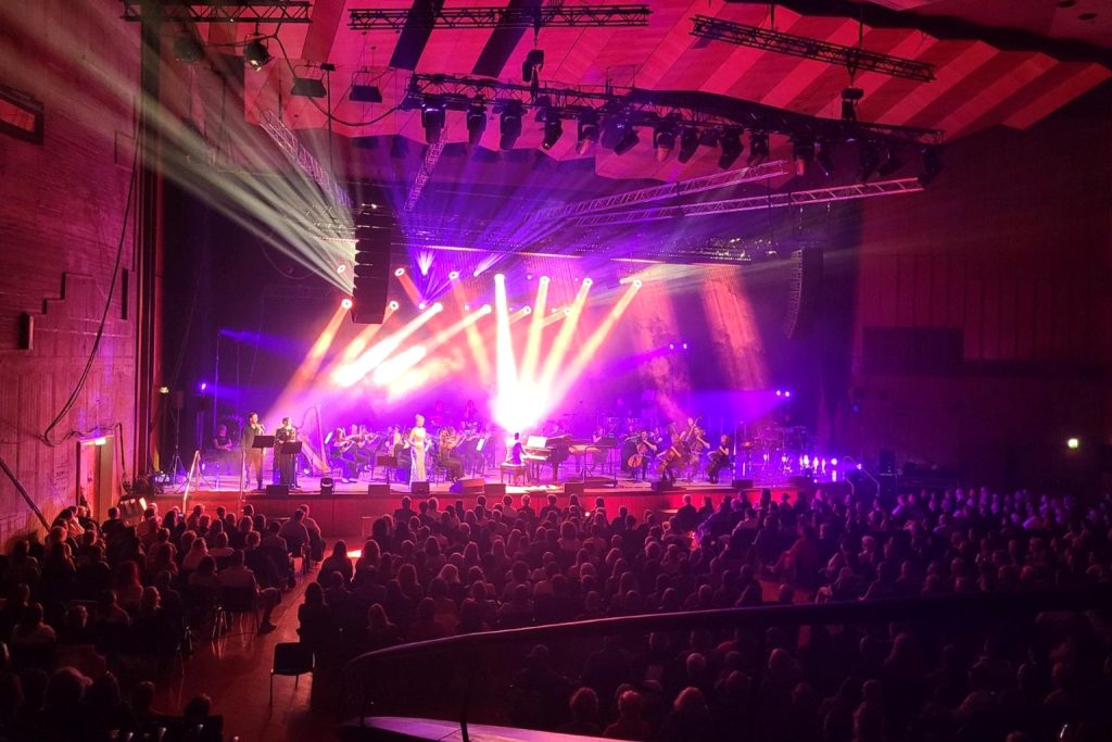 Orchester auf der Bühne bei Kosmos und Orchester in Stuttgart 2026, mit farbigen Lichtstrahlen und Publikum